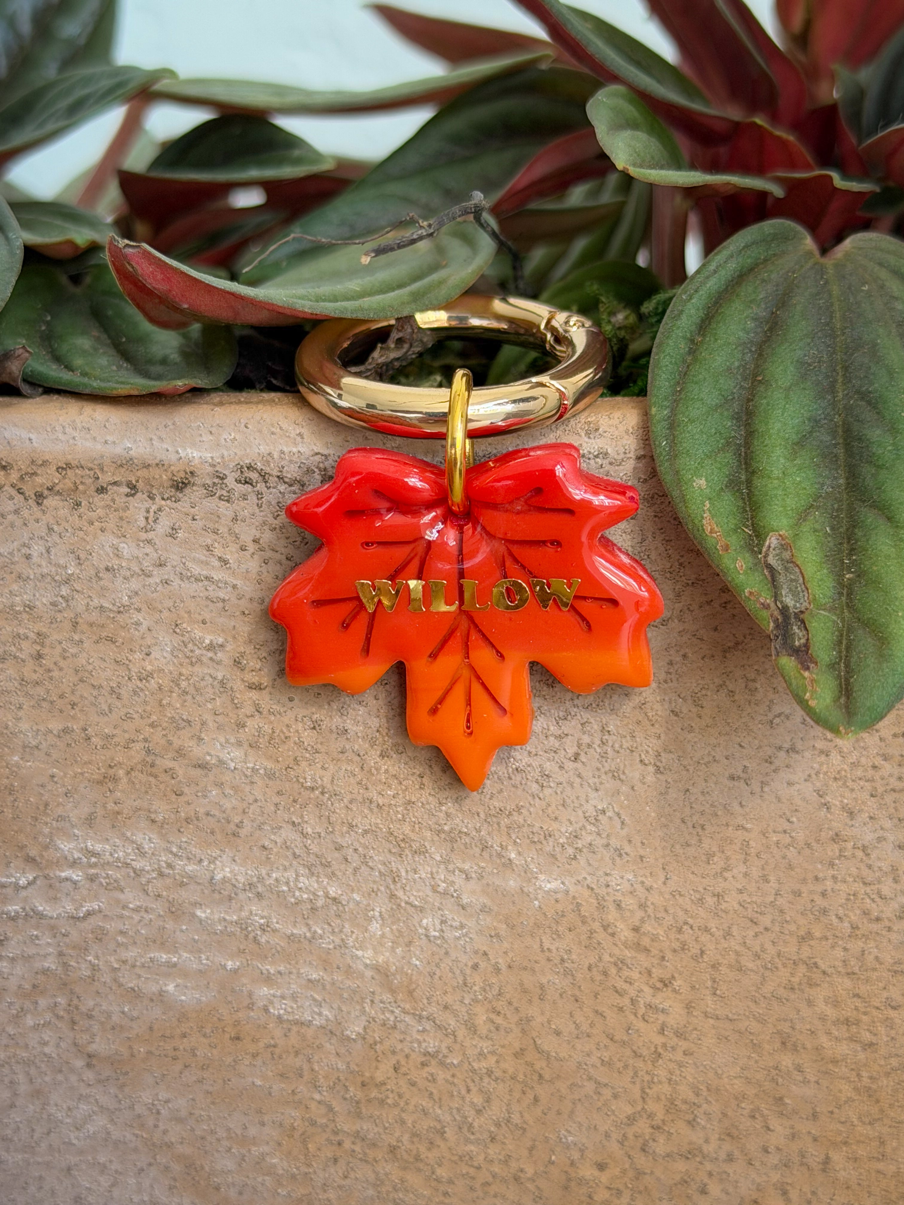 An orange and red maple leaf charm key ring with a gold-toned metal ring and a gold-etched 'WILLOW' brand name, resting on a beige stone planter with green and burgundy foliage.