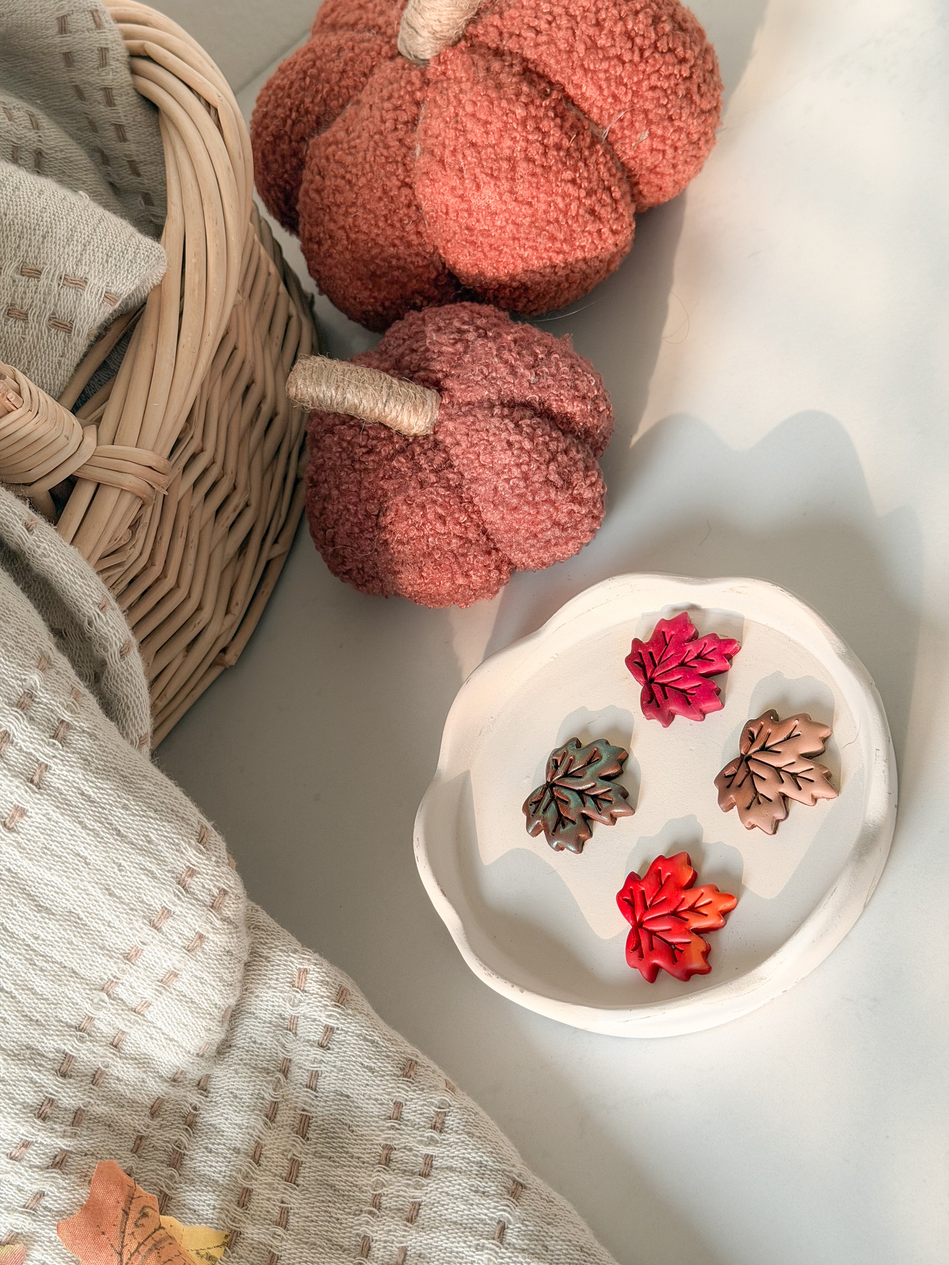 A styled flat-lay featuring a white ceramic dish holding three small dried flower or leaf-like botanical pieces in pink, red-orange, and brown, alongside a wicker basket, textured knit blanket, and two fuzzy terracotta decorative pumpkins on a light surface.