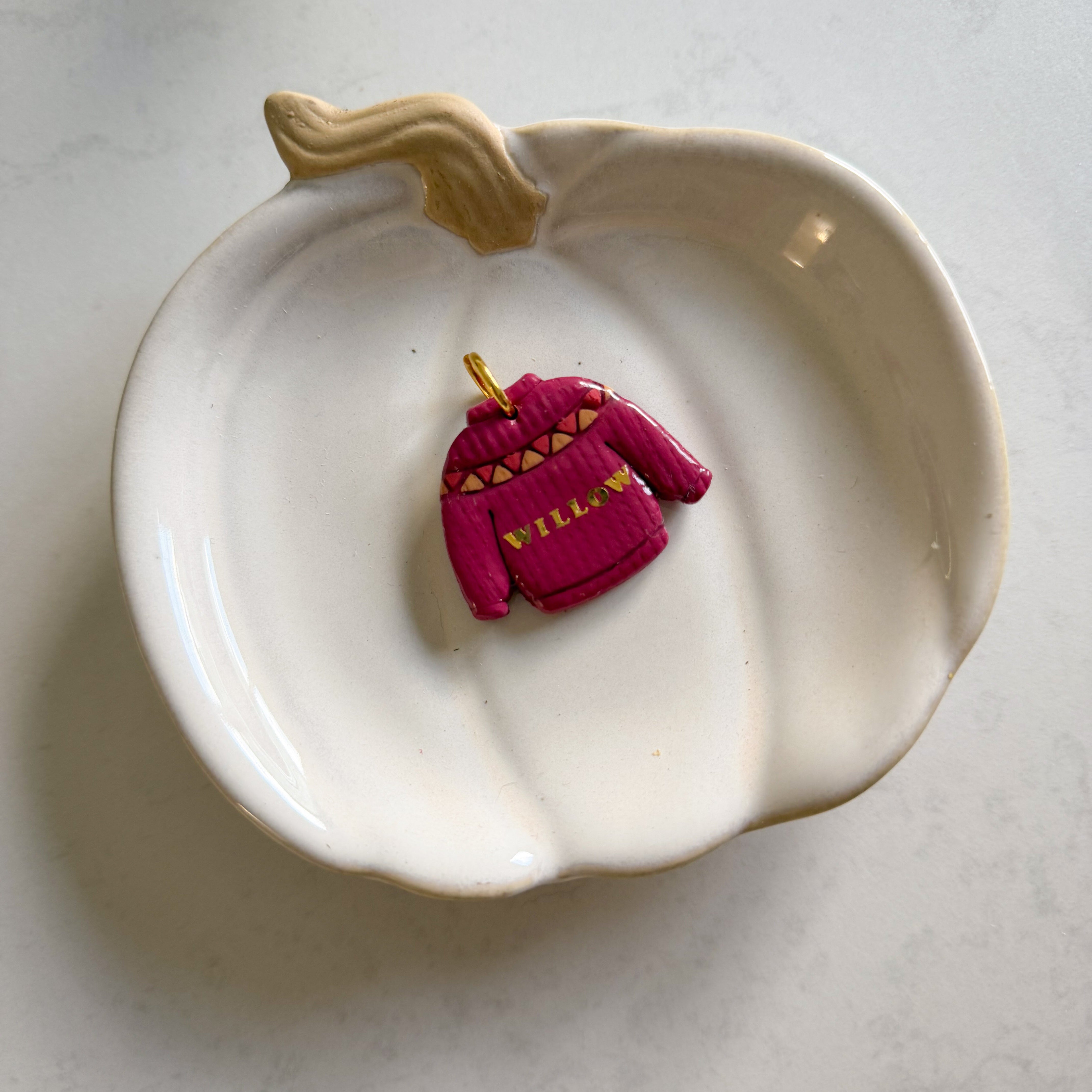 A small pink sweater-shaped jewelry charm with gold lettering and details, displayed inside a white ceramic pumpkin dish on a light gray marble surface.