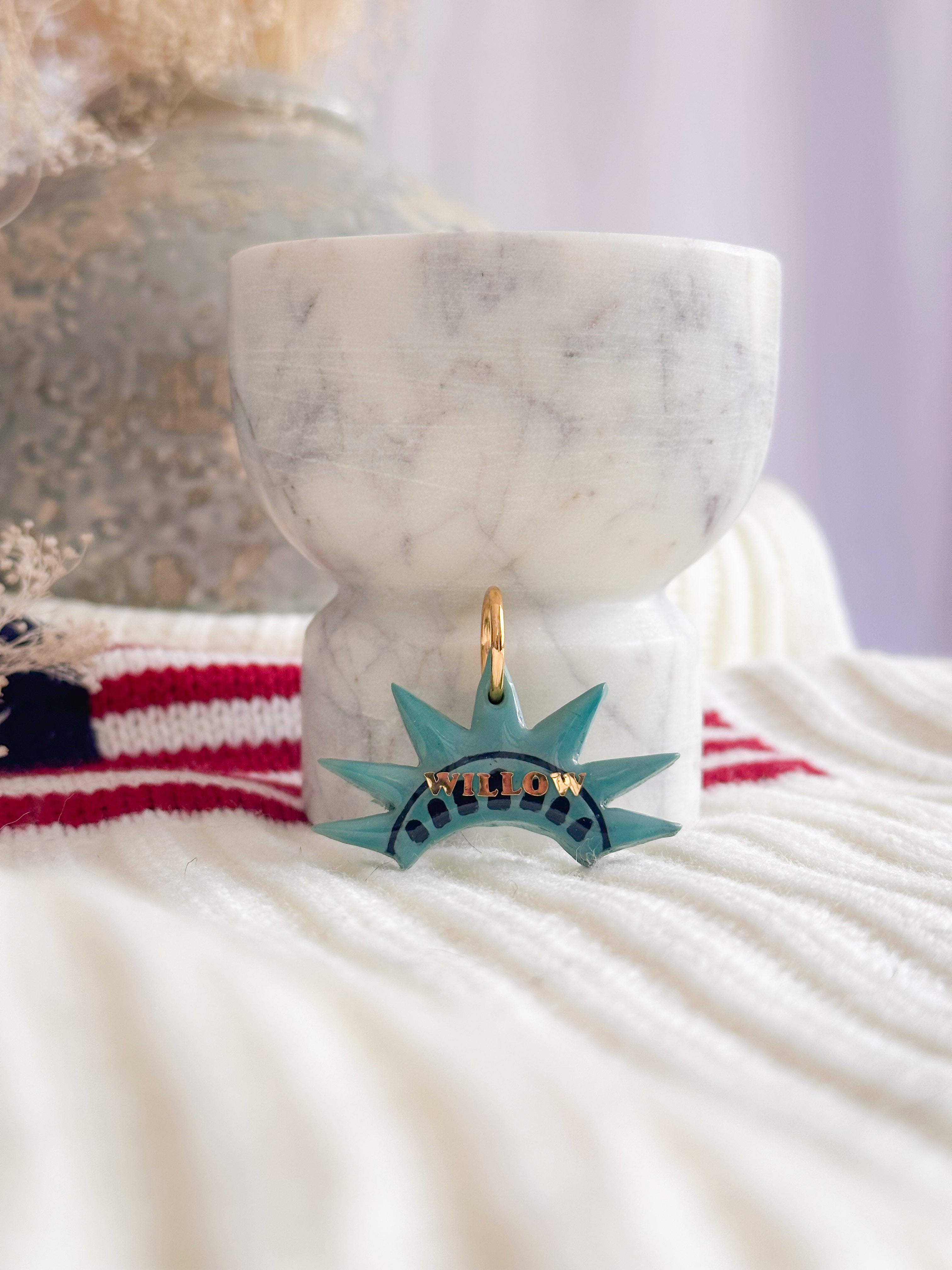 Teal necklace pendant shaped like the Statue of Liberty on a marble surface with an American flag in the background.
